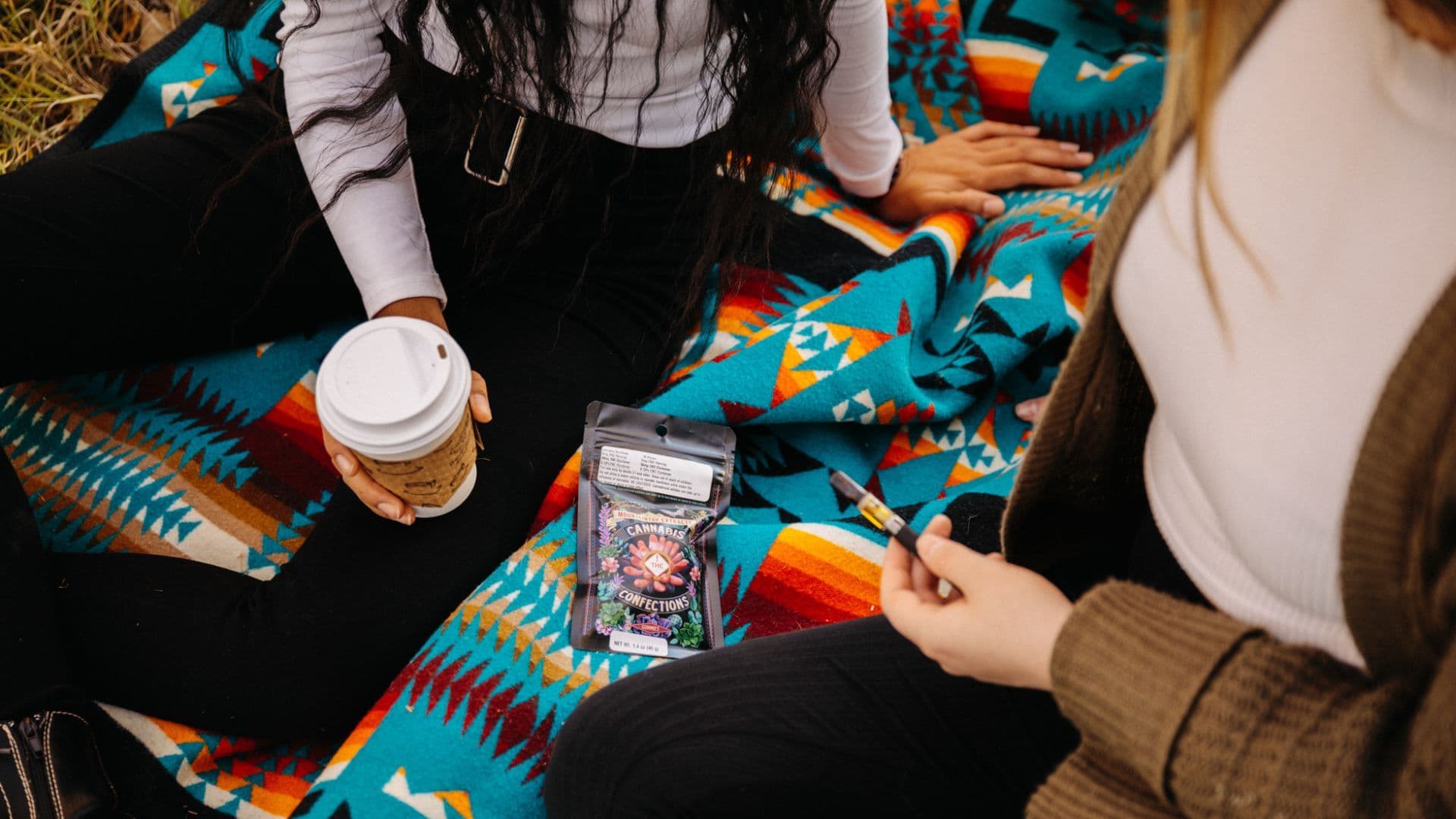 photo of two people having a picnic. you see them from the shoulders down, one is holding a cup of coffee, one is holding a cannabis vape pen. there is a package of edibles from Mountaintop Extracts on the teal blanket.